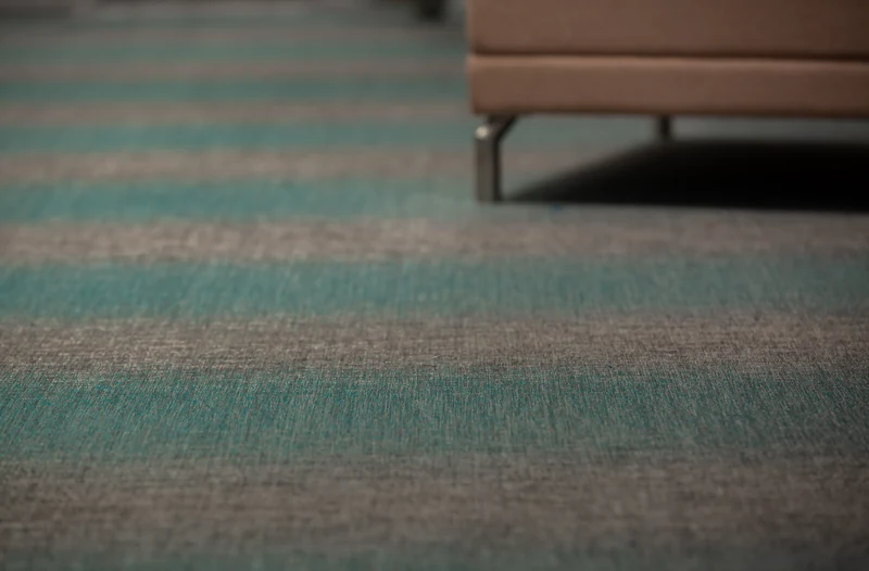 Woven vinyl wall to wall flooring in wide teal and grey stripes, seen close up beside an upholstered bench